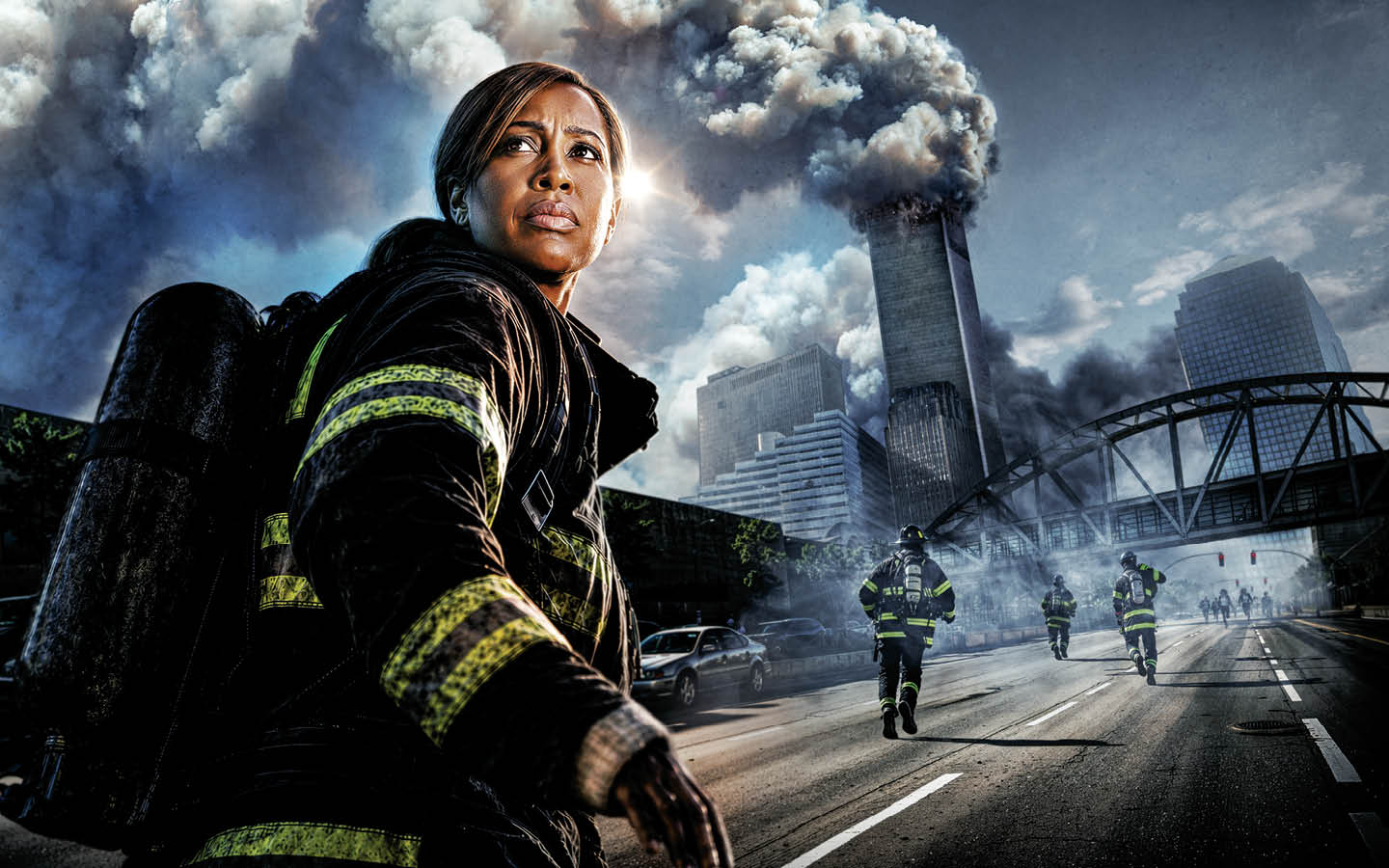 A firefighter in full gear stands on a city street as smoke billows from a tall building behind her and other firefighters run toward it