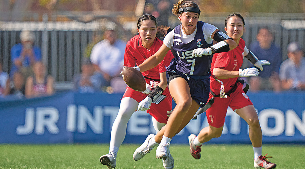 A USA flag football player runs with the ball as two defenders in red pursue her