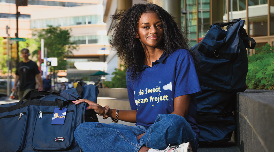 A young woman sits outdoors with duffel bags, wearing a Sweet Dream Project shirt